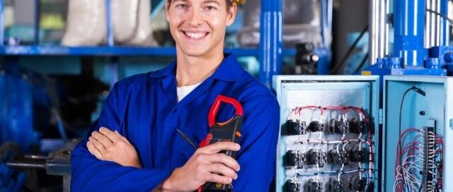 Smiling local electrician holding a multimeter beside an open electrical panel in Prescott Valley advising on How To Choose The Best Local Electrician Near Me